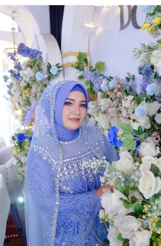 Pengantin berkerudung biru berdiri di antara rangkaian bunga putih dan biru.