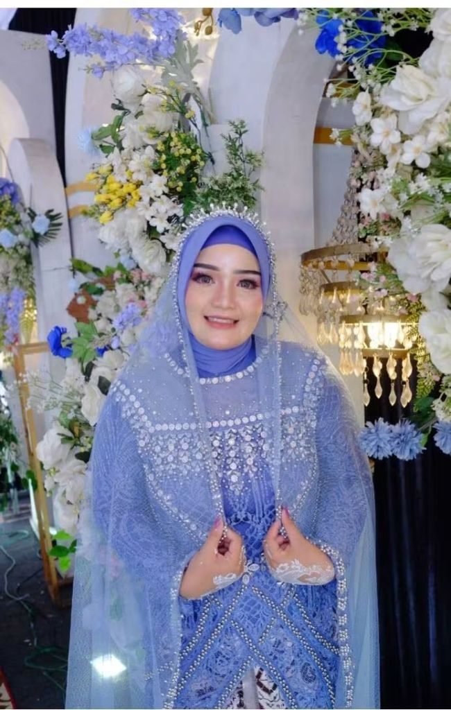 Pengantin mengenakan gaun dan kerudung biru bertabur mutiara di depan dekorasi bunga.