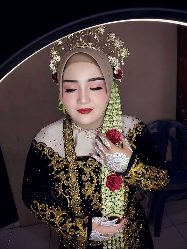 Pengantin menutup mata dengan tenang, mengenakan mahkota emas bertabur bunga dan kebaya hitam bersulam emas.