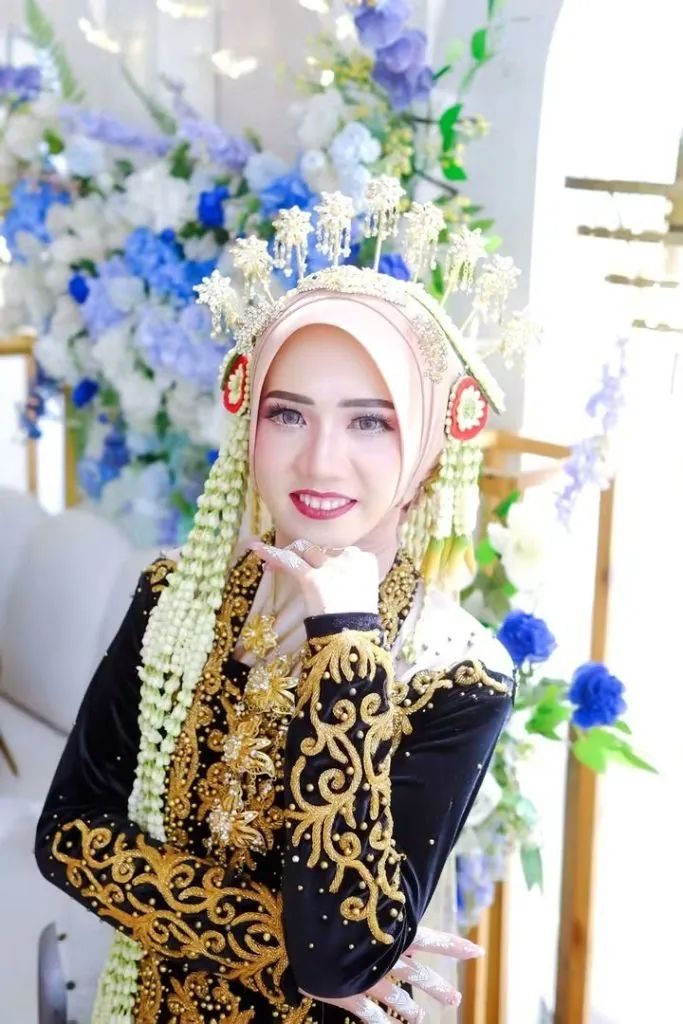 Pengantin dengan hiasan kepala emas mengangkat tangan berhias henna di samping wajah.