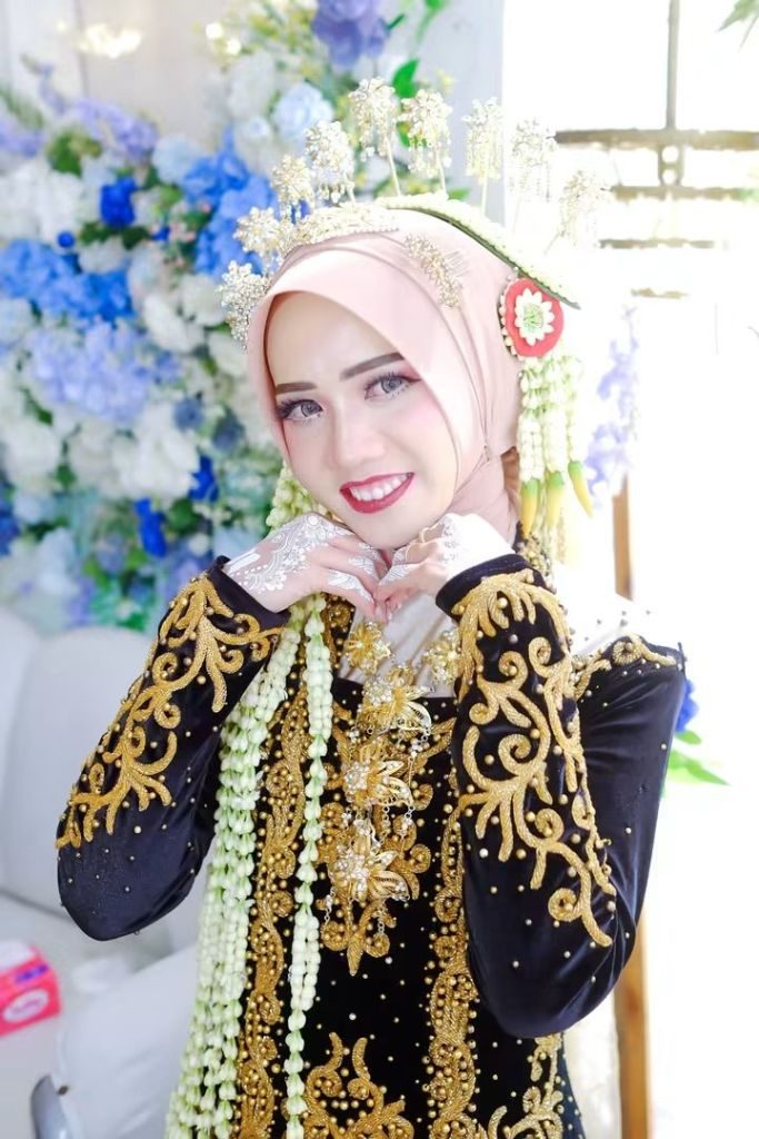 Pengantin tersenyum dengan kedua tangan berhias henna di depan dada, mengenakan kebaya hitam bersulam emas.