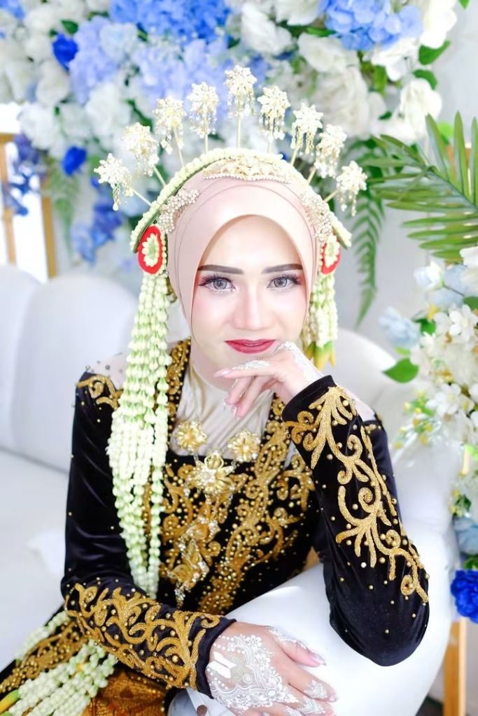 Pengantin dengan riasan paes, kebaya hitam bersulam emas, dan rangkaian melati, duduk di depan dekorasi bunga.