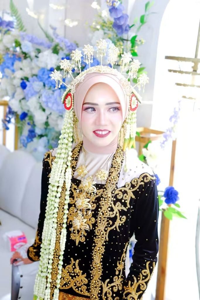 Pengantin berdiri mengenakan kebaya hitam bersulam emas dengan rangkaian melati di kedua sisi.