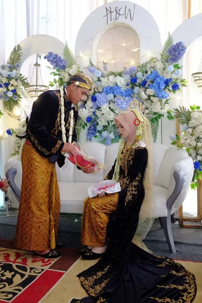 Prosesi pernikahan: pengantin pria berdiri membawa seserahan di hadapan pengantin perempuan yang duduk.