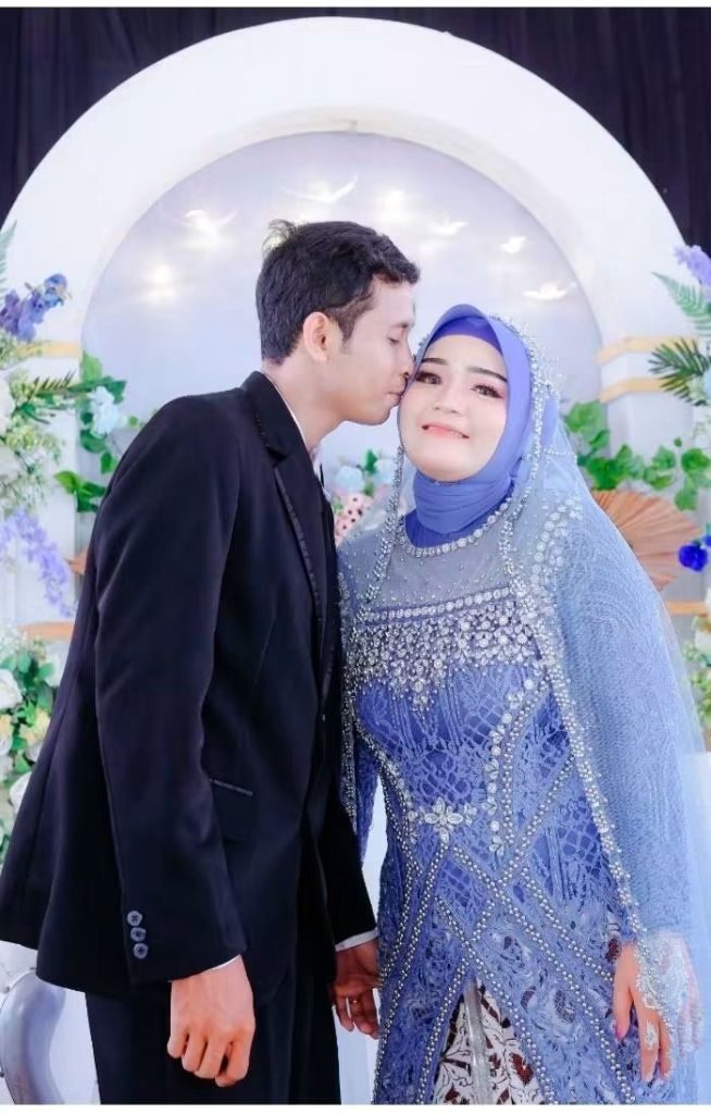 Pengantin pria berjas hitam mencium pipi pengantin perempuan berbusana biru bertabur mutiara.