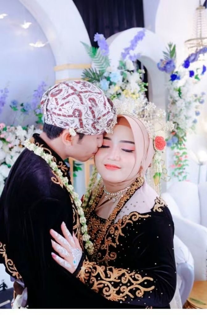 Pengantin pria mencium dahi pengantin perempuan yang memejamkan mata, di depan dekorasi bunga.