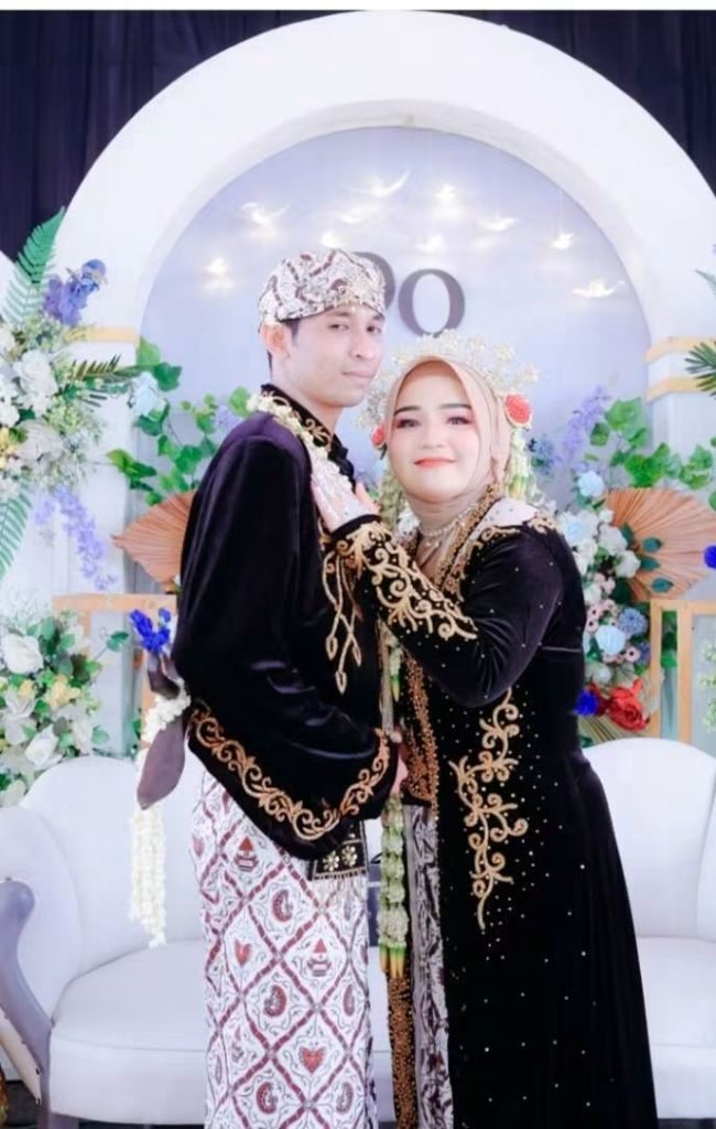 Pasangan pengantin berdiri berdampingan dengan busana adat hitam bersulam emas di pelaminan.