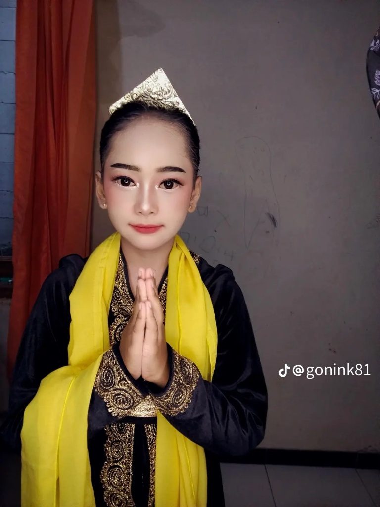 Riasan panggung tradisional dengan hiasan kepala emas dan selendang kuning, tangan menyatu memberi salam.